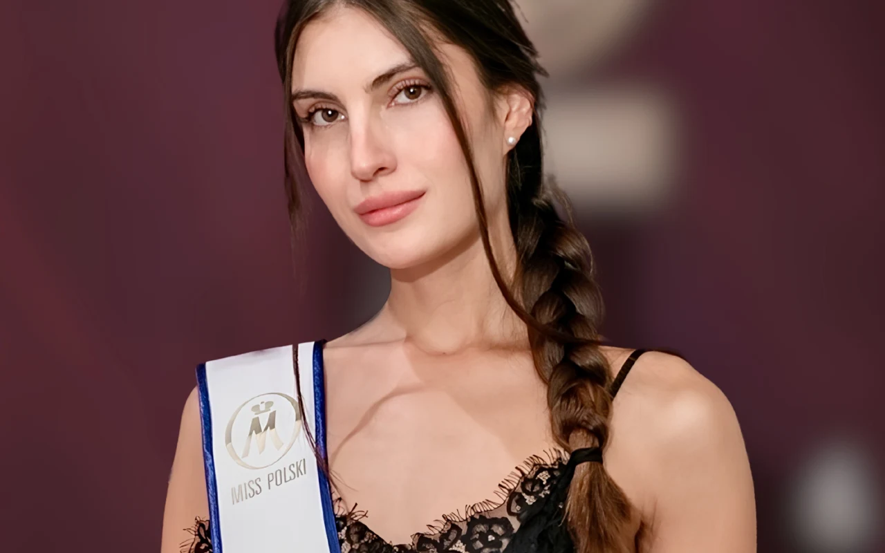 Młoda kobieta z długim warkoczem, ubrane w czarną koronkową sukienkę na ramiączkach, nosi białą szarfę z napisem „Miss Polski” na tle fioletowego tła.