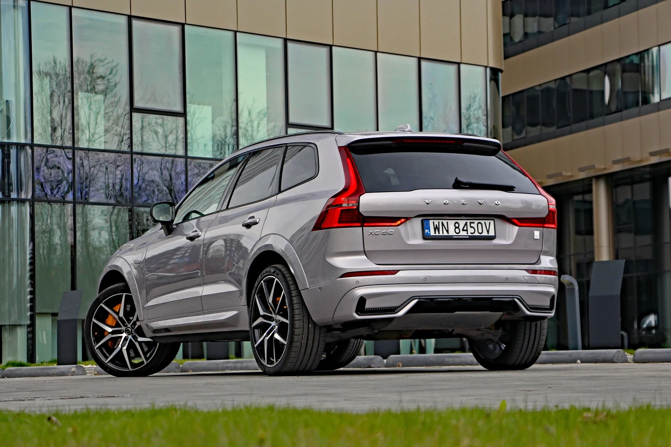 Srebrny SUV Volvo na dużych, sportowych felgach stoi zaparkowany przed nowoczesnym budynkiem o szklanej elewacji.