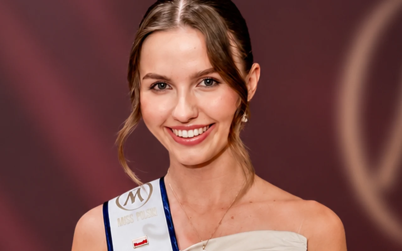 Uśmiechnięta młoda kobieta z jasnymi włosami upiętymi do tyłu, ubrana w elegancką suknię i szarfę z logo oraz małą flagą Polski, pozująca na tle w kolorze bordowym.