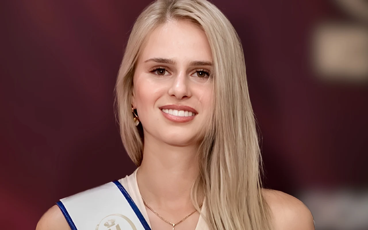Uśmiechnięta młoda kobieta o długich blond włosach, ubrana w elegancką szarfę z ozdobnym medalionem, pozująca na tle w odcieniach bordo.