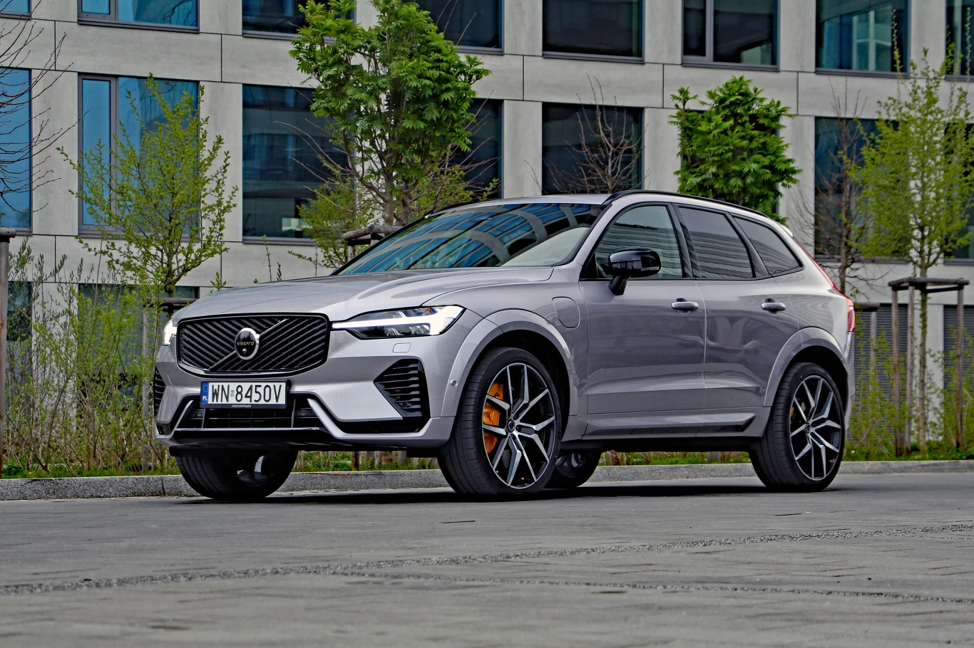 Srebrny SUV marki Volvo z dużymi felgami i sportowymi detalami stoi zaparkowany przy nowoczesnym budynku, otoczony skromną zielenią.