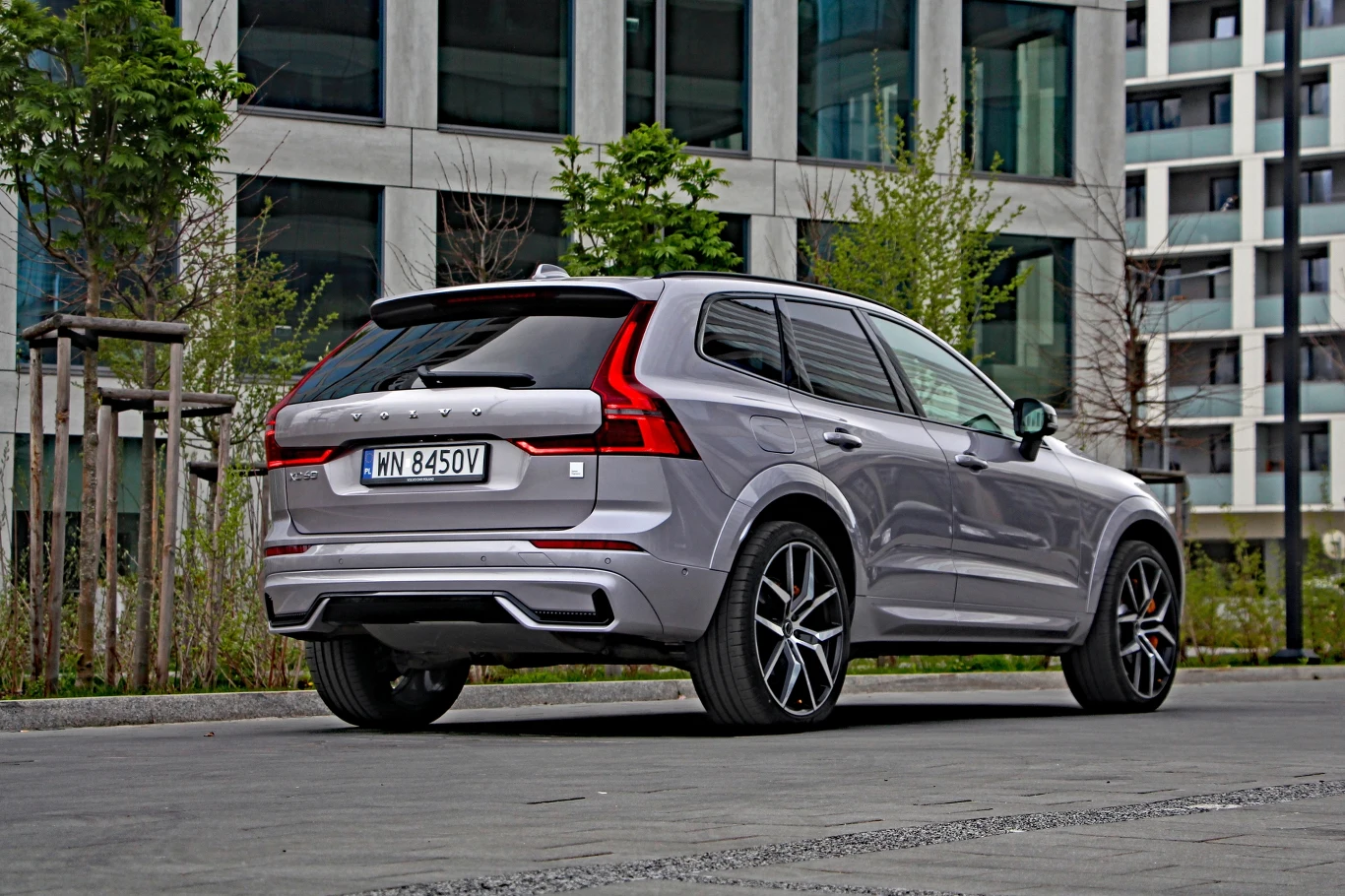 Srebrny SUV Volvo XC60 zaparkowany na tle nowoczesnych budynków ze szklaną fasadą, otoczony niewielkimi drzewami i miejskim chodnikiem.
