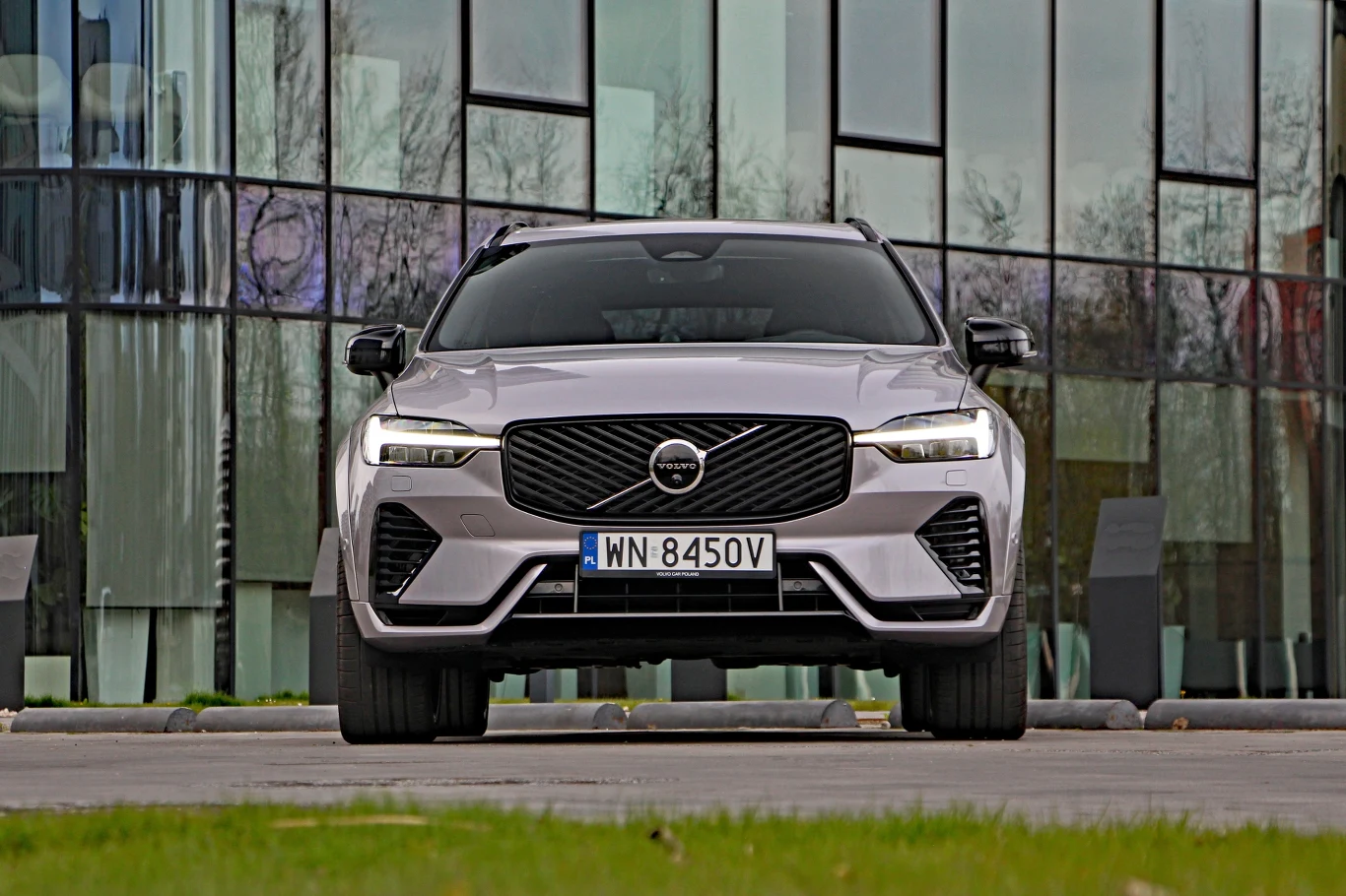 Srebrny SUV marki Volvo o nowoczesnym designie, ustawiony przodem na tle przeszklonego budynku, światła LED i charakterystyczna osłona chłodnicy podkreślają dynamiczny wygląd samochodu.