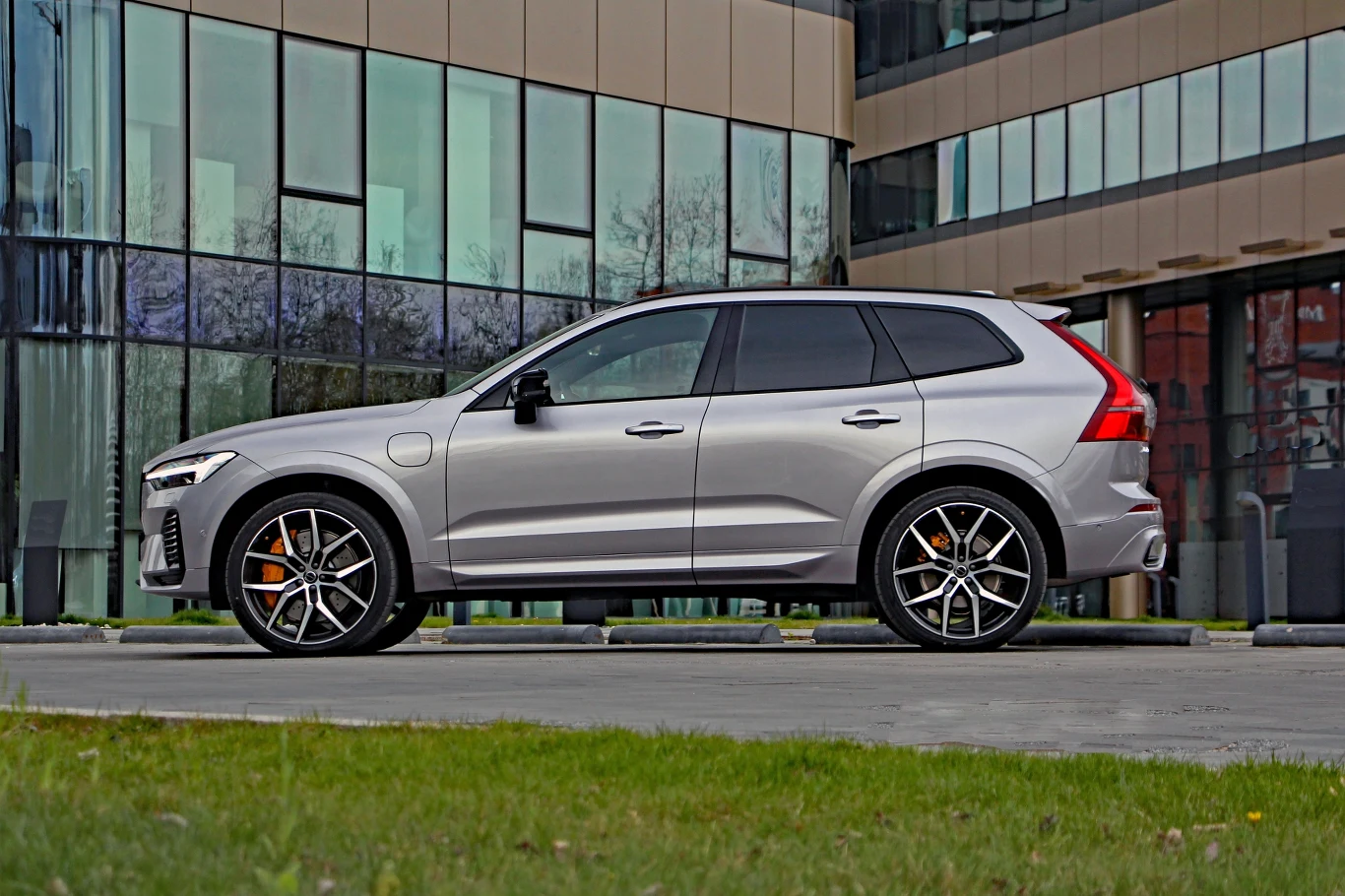 Srebrny SUV o nowoczesnych liniach nadwozia, duże sportowe felgi z pomarańczowymi zaciskami hamulców, panoramiczne okna budynku biurowego w tle.
