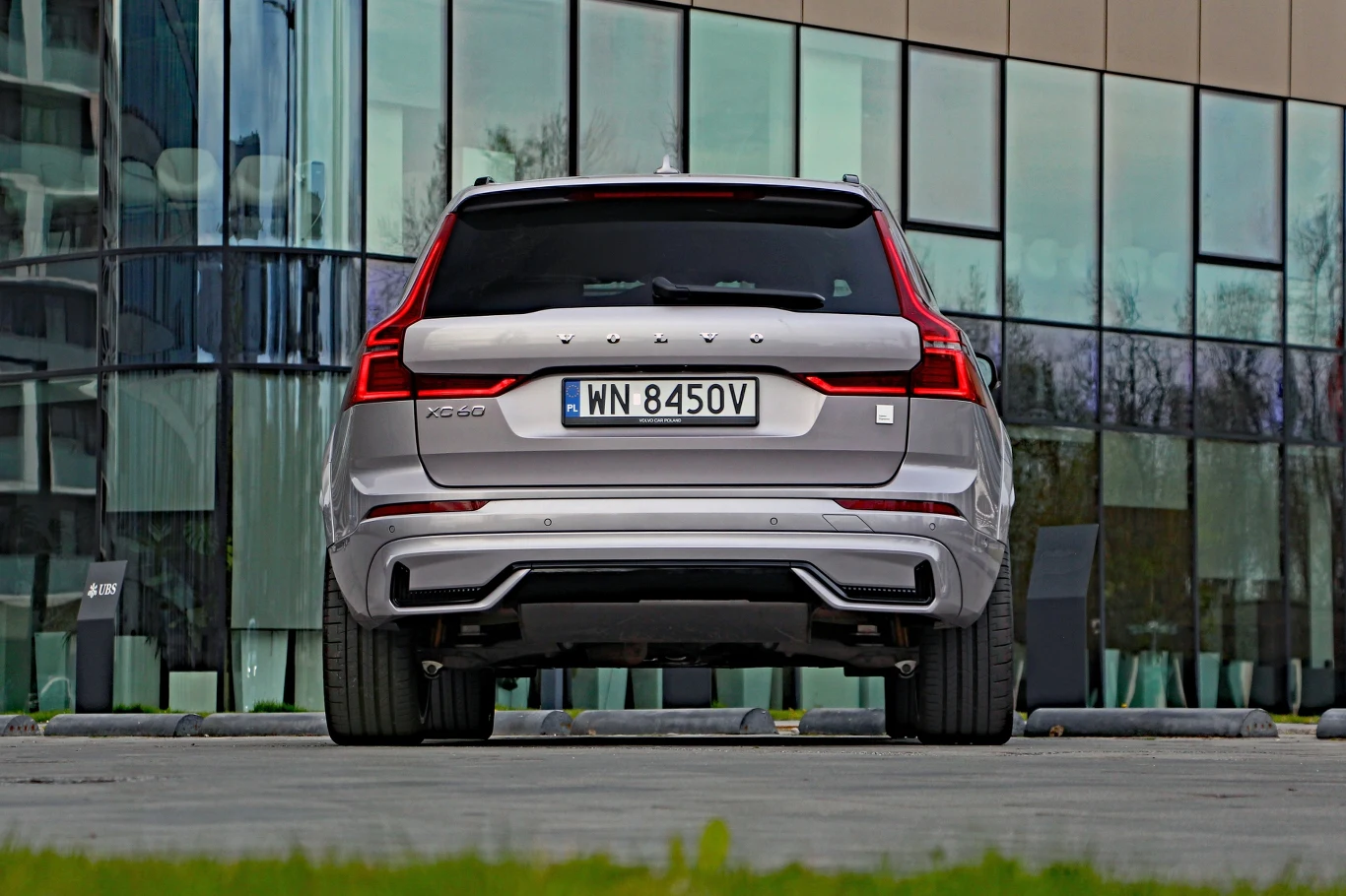 Srebrny Volvo XC60 zaparkowany tyłem na tle przeszklonego budynku, widoczne charakterystyczne tylne światła LED oraz tablica rejestracyjna.