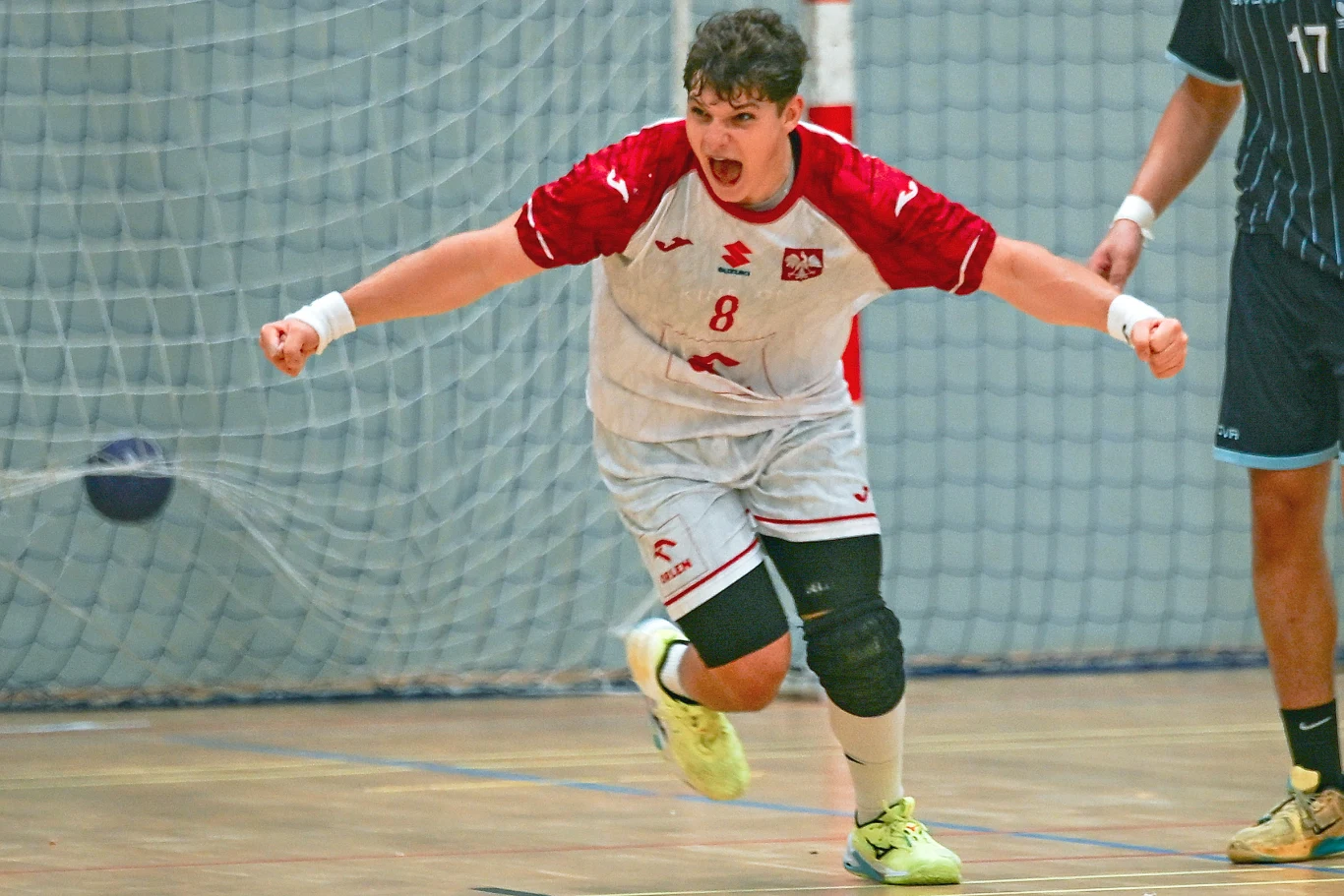Zawodnik w białoczerwonym stroju z numerem 8 świętuje zdobycie bramki podczas meczu piłki ręcznej, tuż przy bramce na hali sportowej.