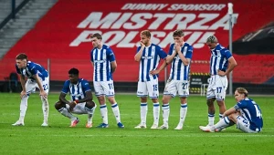Siedmiu piłkarzy w biało-niebieskich strojach z wyraźnie rozczarowanymi minami stoi i siedzi na murawie stadionu, na tle czerwonego baneru z napisem 'Mistrza Polski'.