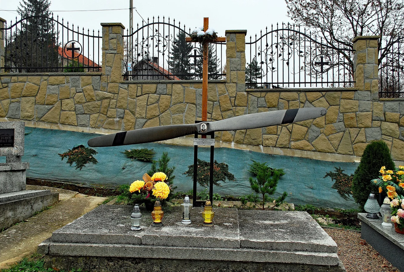 Nagrobek z postawionym dużym śmigłem samolotu oraz krzyżem, otoczony kwiatami i zniczami, na tle kamiennego muru z metalowym ogrodzeniem.