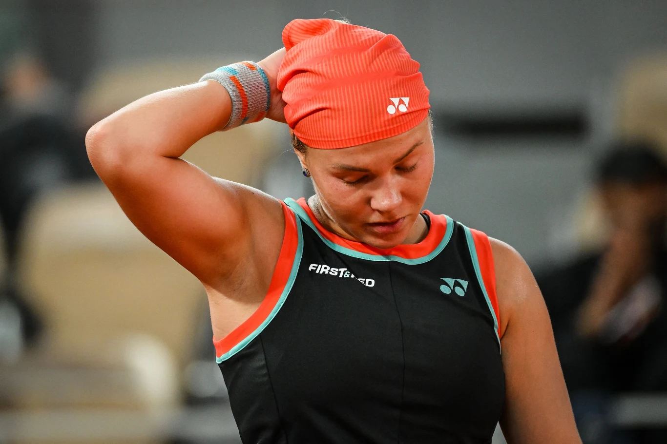 Zawodniczka tenisowa w czarno-czerwonym stroju sportowym oraz opasce, z opuszczoną głową i ręką uniesioną do tyłu szyi, wyrażająca zmęczenie lub zamyślenie.