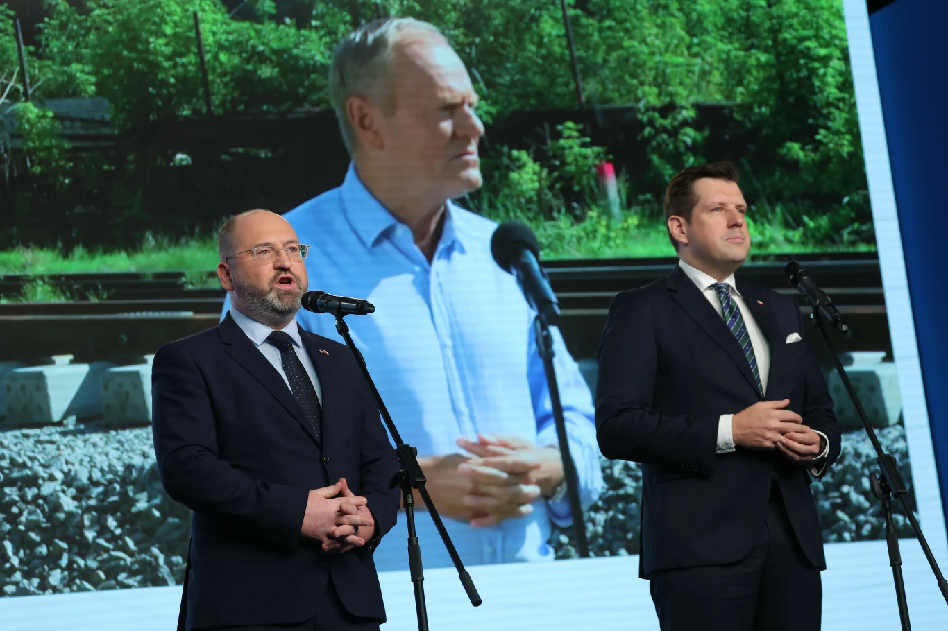 Dwaj mężczyźni w garniturach przemawiają przy mikrofonach, w tle duży ekran prezentujący zbliżenie mężczyzny w jasnej koszuli na tle torów kolejowych i zieleni.