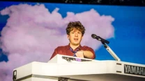 Mężczyzna grający na białym keyboardzie marki Yamaha, za nim scenografia z chmurami na niebieskim tle, po bokach mikrofon na statywie.