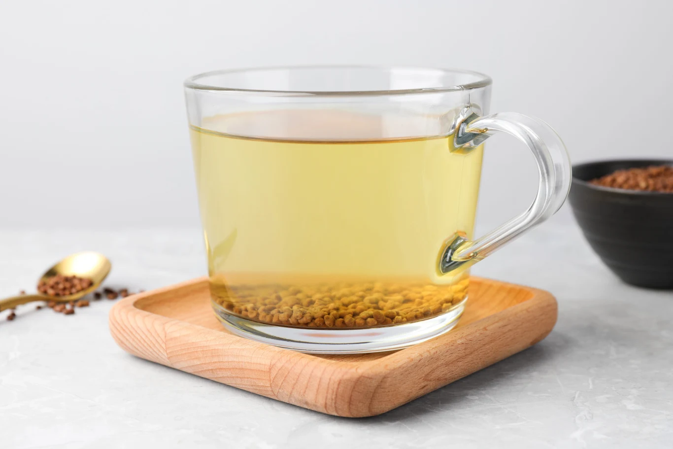 Ziarna, z których parzy się herbatę, zaliczane są do tzw. super foods. Szklanka jasnego naparu z prażonych ziaren gryki na drewnianej podstawce, obok łyżeczka i miseczka z ziarnami.