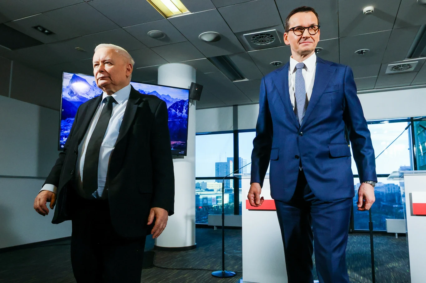 Prezes PiS-u Jarosław Kaczyński i były premier Mateusz Morawiecki Dwóch mężczyzn w garniturach stoi w nowoczesnym wnętrzu biurowym z dużymi oknami i ekranami w tle.