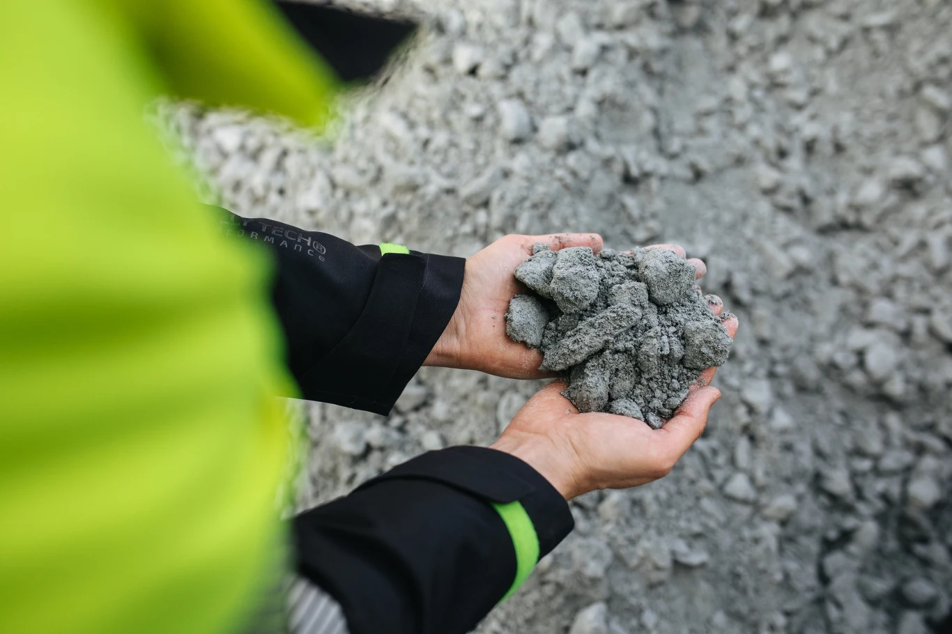 Dłonie w czarno-zielonych rękawach trzymające grudki szarego materiału przypominającego kruszywo lub cement.