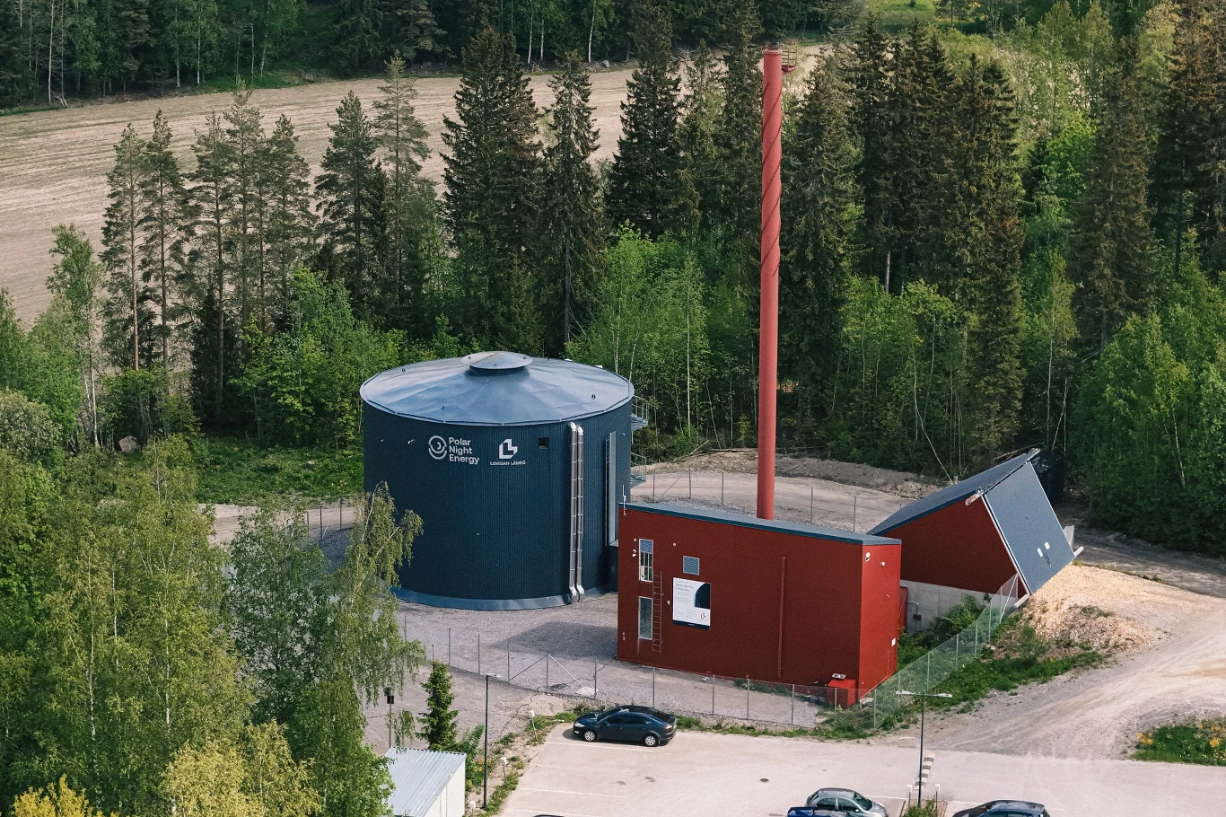 Bateria piaskowa w Porainen Nowoczesna kotłownia z dużym, cylindrycznym zbiornikiem na wodę oraz wysokim, czerwonym kominem, otoczona ogrodzeniem i położona wśród drzew na skraju lasu.