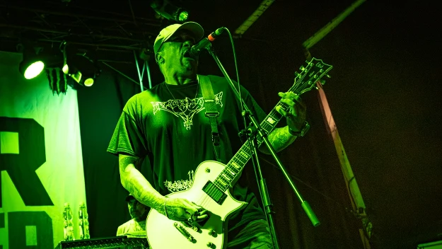 Mężczyzna w czapce z daszkiem gra na białej gitarze elektrycznej i śpiewa do mikrofonu podczas koncertu, scena oświetlona zielonym światłem.