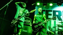 Dwóch muzyków w dynamicznej pozie na scenie podczas koncertu, jeden gra na gitarze basowej, drugi na gitarze elektrycznej, wokół nich profesjonalny sprzęt muzyczny i intensywne zielone oświetlenie sceniczne.