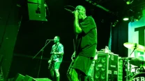 Wokalista i gitarzysta zespołu hardcore występują na scenie, otoczeni zielonym światłem, w tle widoczna perkusja oraz kolumna głośnikowa z naklejkami.