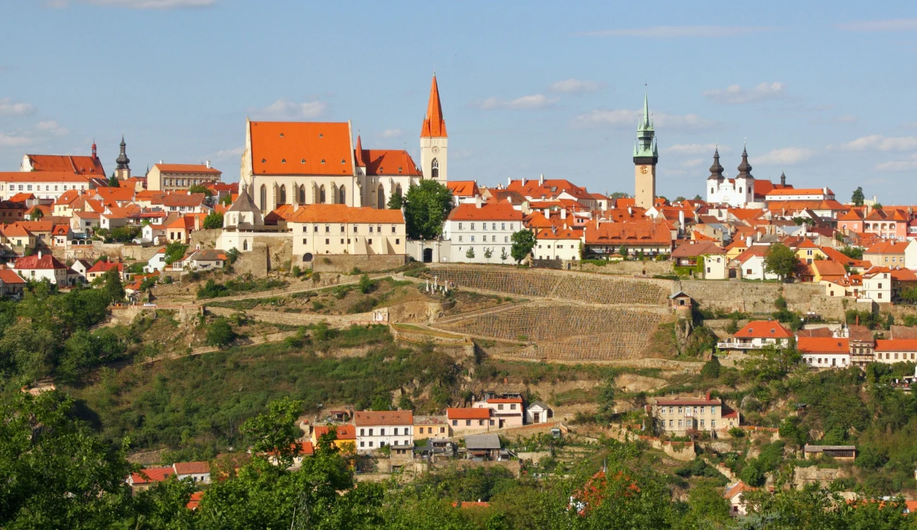 Co warto zobaczyć w Znojmo? Panorama Znojmo z kościołem o czerwonym dachu, wysoką wieżą zegarową i zabudową otoczoną zielenią i wzgórzami.