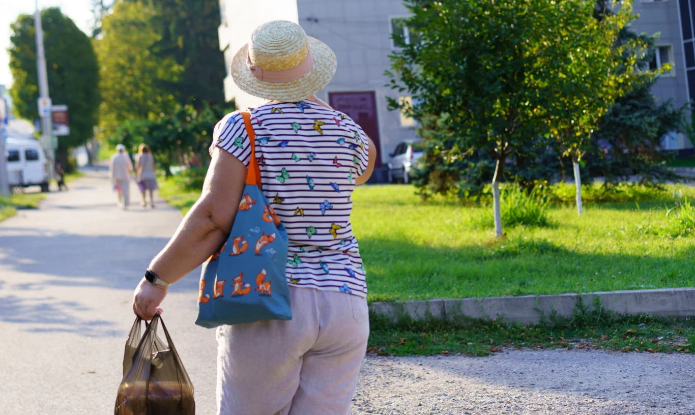 Kobieta w kapeluszu i kolorowej koszulce z torbą na ramieniu oraz reklamówką w ręce idzie chodnikiem w stronę zielonego skwerku przy budynku mieszkalnym.