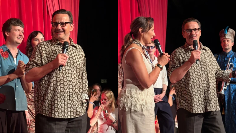 Mężczyzna w okularach trzyma mikrofon i przemawia przed publicznością, wokół niego stoją inne osoby, a za nimi widoczna jest czerwona kotara teatralna.