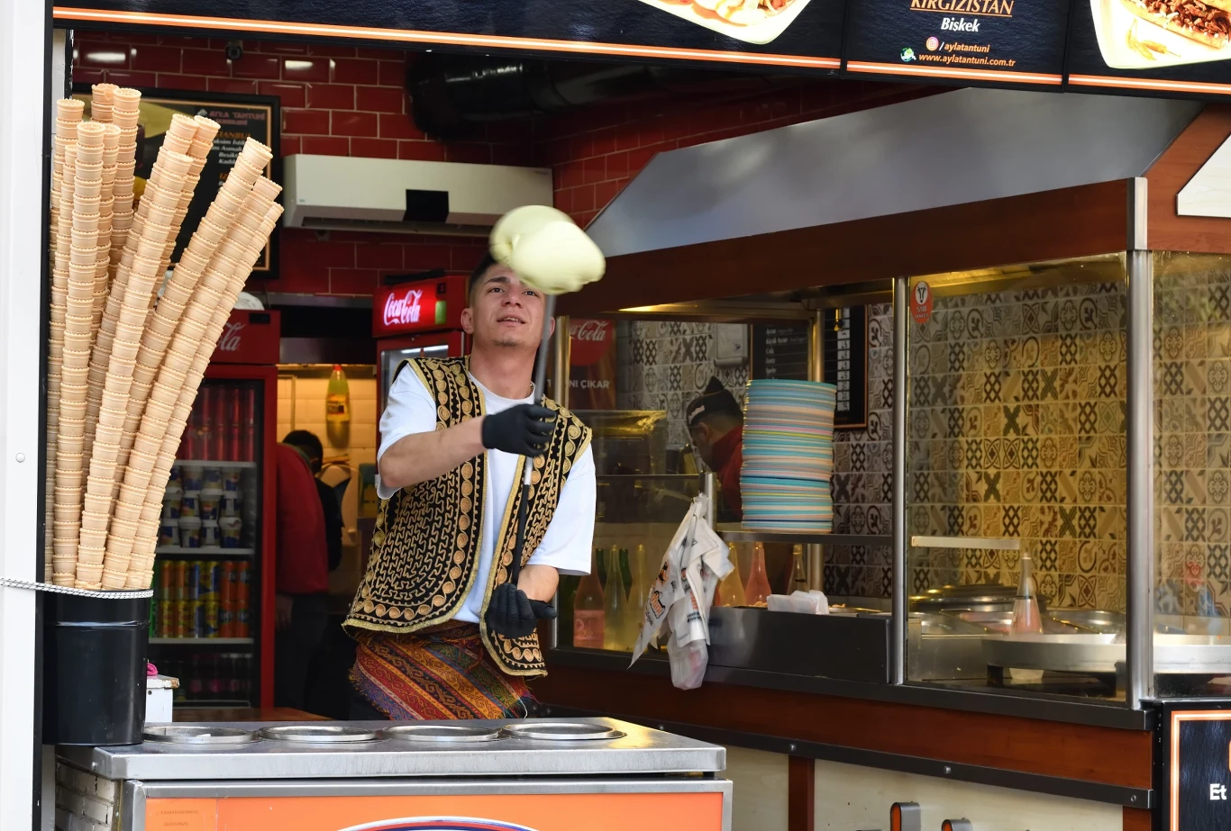Turecki show z gałką lodów na długim kijku może nas słono kosztować Sprzedawca lodów w tradycyjnym stroju wykonuje widowiskową sztuczkę z porcją lodów nad ladą, obok ustawiony stojak z waflami do lodów.