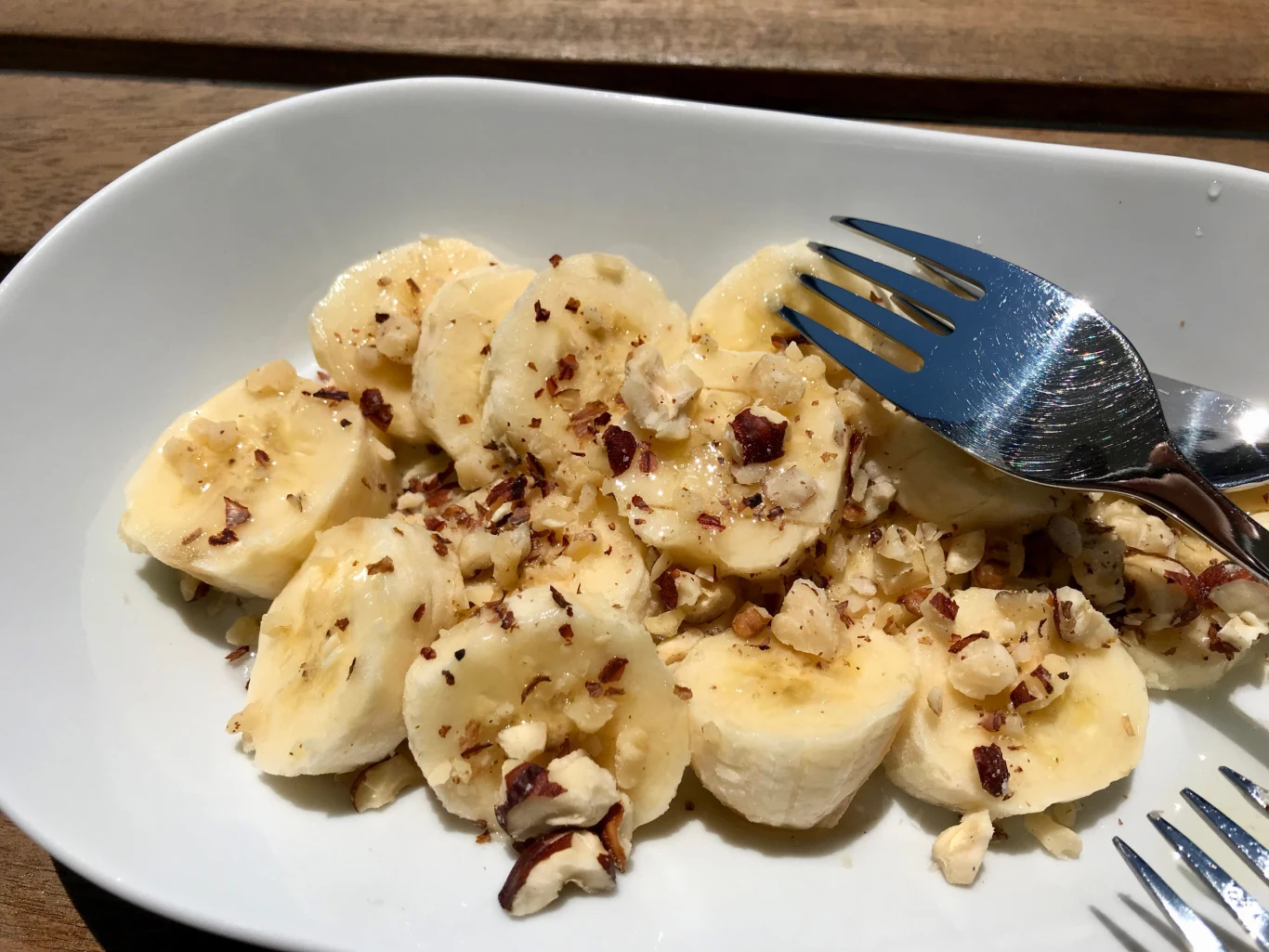 Banany to dobre źródło potasu, który pomaga zbić zbyt wysokie ciśnienie krwi Plasterki banana posypane pokruszonymi orzechami na białym talerzu, obok widelce.
