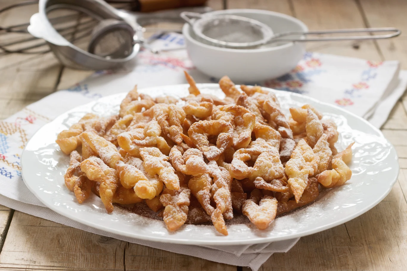 Faworki zachwycają delikatnym ciastem i charakterystyczną chrupkością. Talerz z faworkami posypanymi cukrem pudrem na drewnianym stole, obok sitko i biała miseczka.