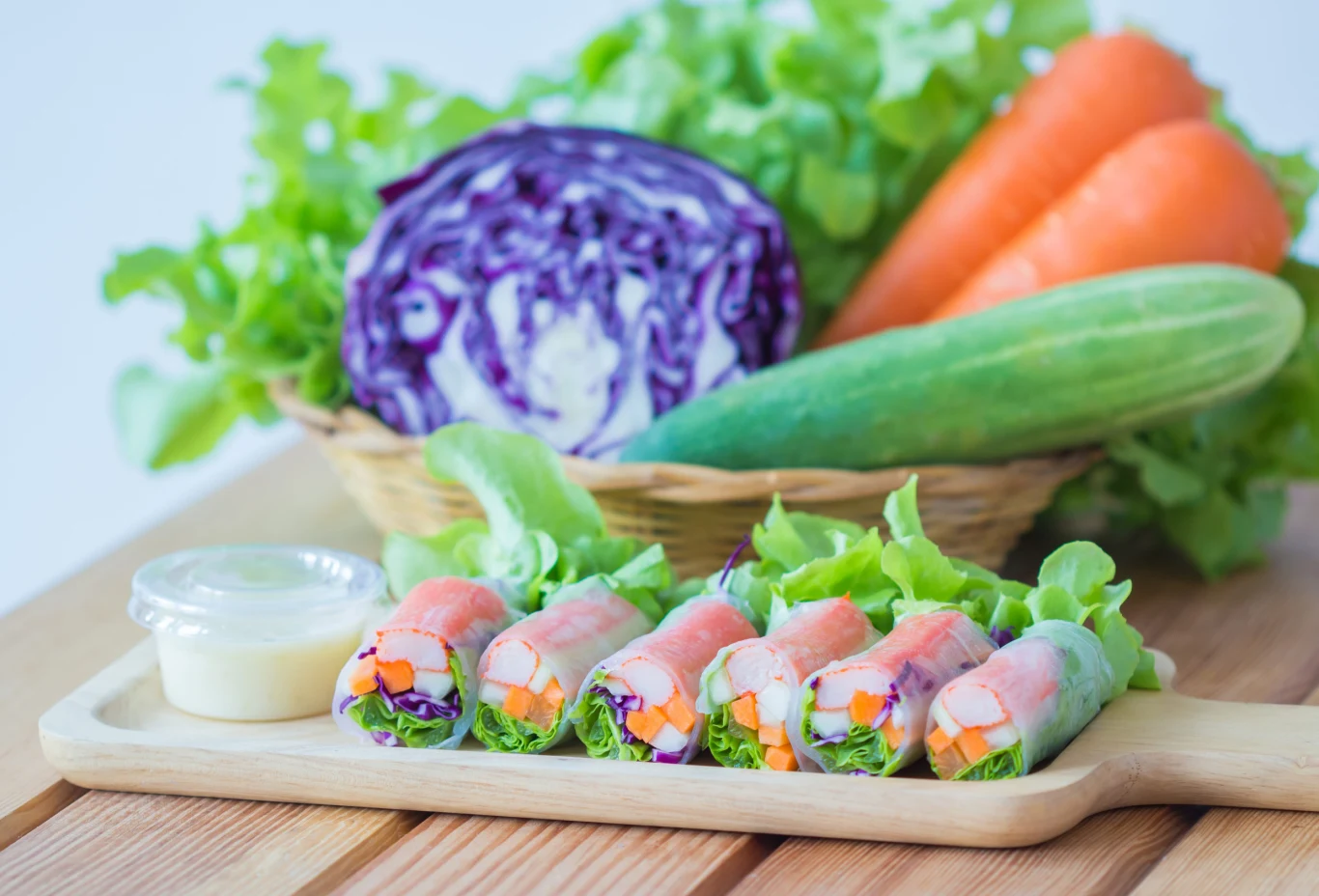 Spring rolls – lekkie wietnamskie roladki pełne świeżych warzyw i dodatków. Kolorowe spring rollsy z warzywami na drewnianej desce obok sosu, w tle sałata, czerwona kapusta, marchewki i ogórek.