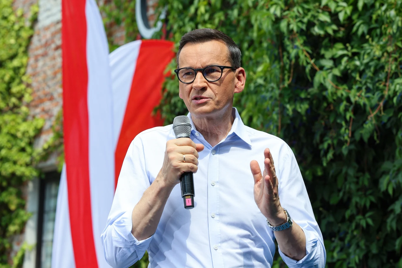 Mateusz Morawiecki zaingurował prace stowarzyszenia Rozwój Plus Mateusz Morawiecki w okularach i białej koszuli mówi do mikrofonu na tle biało-czerwonej flagi i zieleni.