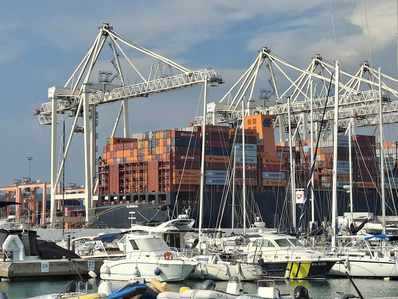 Port w Koprze Ogromny kontenerowiec załadowany kolorowymi kontenerami, obsługiwany przez wysokie dźwigi portowe, w tle liczne jachty zacumowane w marinie.