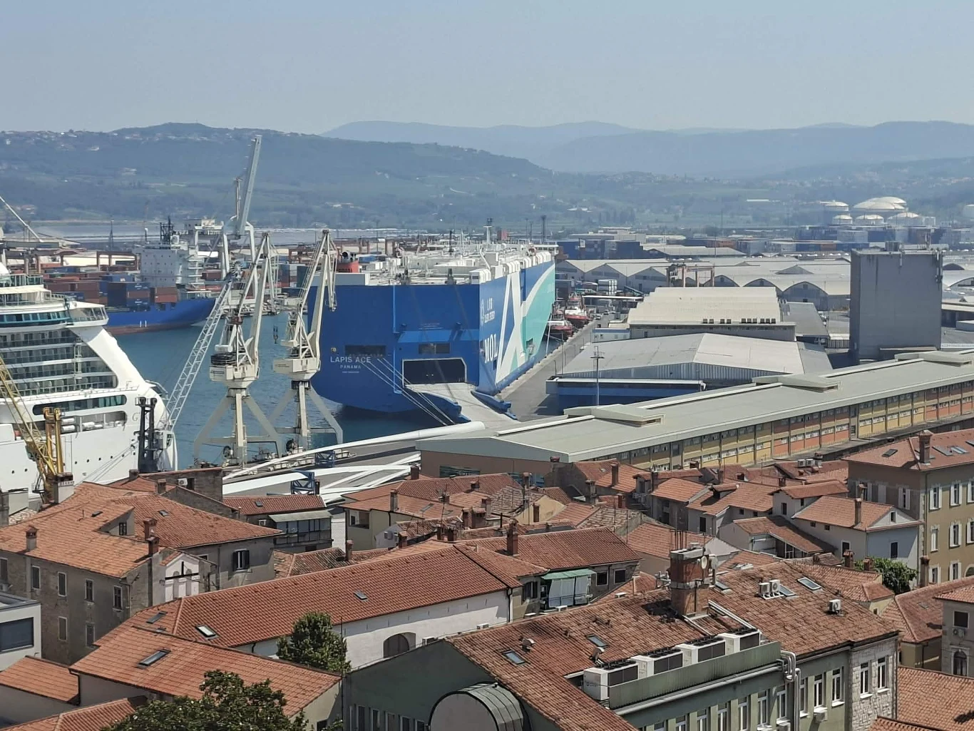 Koperski port widoczny z wieży lokalnego kościoła Olbrzymi statek towarowy zacumowany przy nabrzeżu portowym, wokół którego ustawione są dźwigi portowe i hale magazynowe, na pierwszym planie zabudowa z czerwonymi dachami.