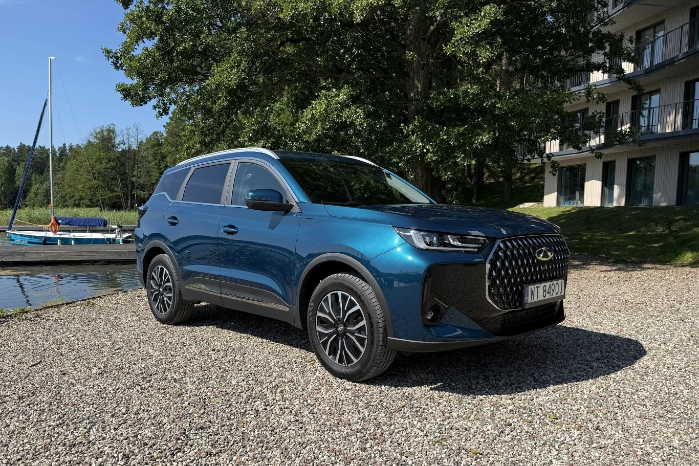 Granatowy SUV z nowoczesnym grillem zaparkowany na żwirowym podłożu obok jeziora i żaglówki, w tle drzewa oraz fragment nowoczesnego budynku.