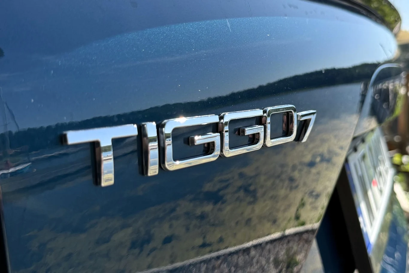 Chromowany emblemat Tiggo 7 na granatowej karoserii samochodu, odbijający otoczenie w lśniącej powierzchni lakieru.