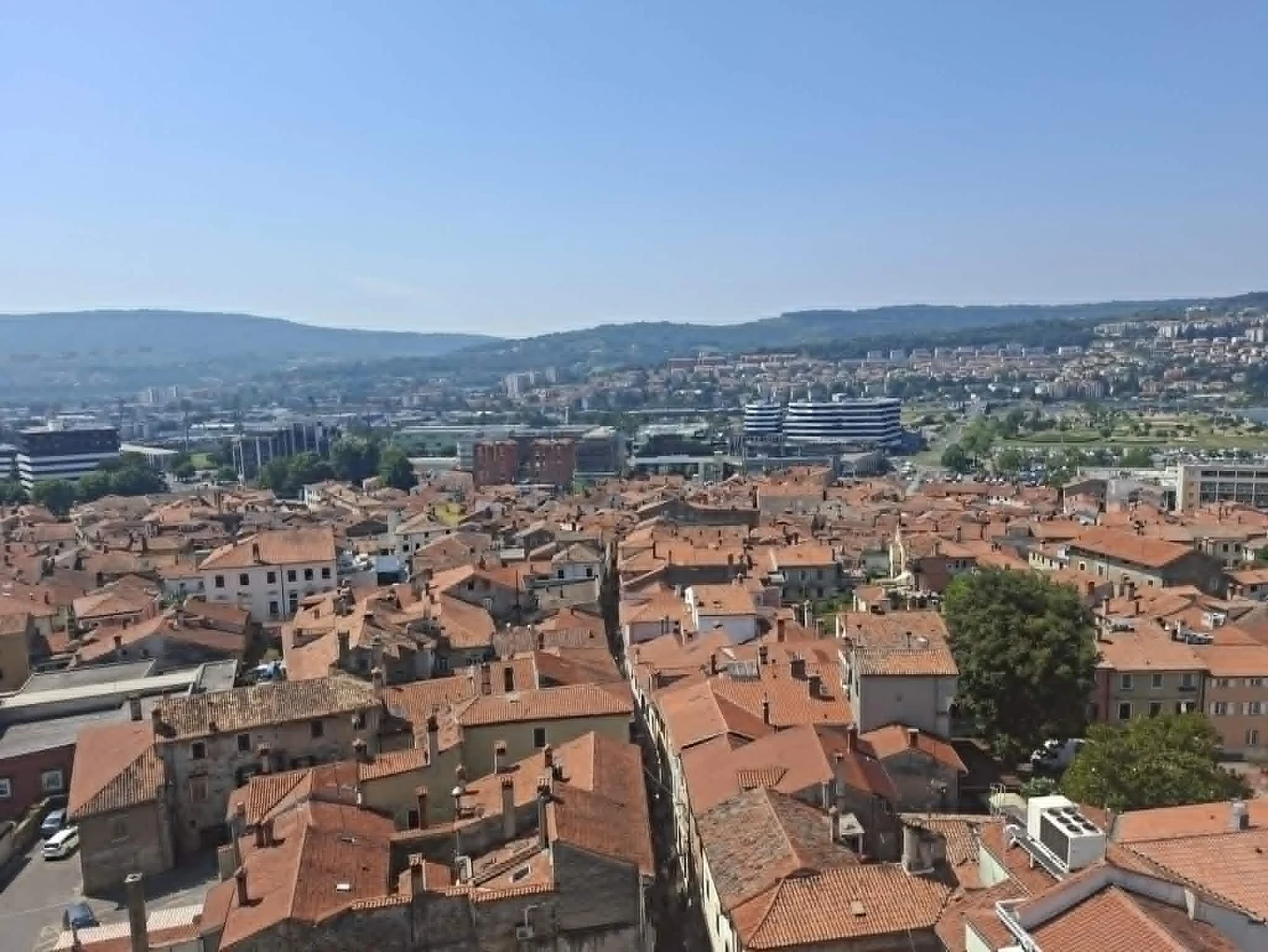 Panorama Kopru i okolic Panorama miasta z niską zabudową o czerwonych dachach, otoczona zielonymi wzgórzami oraz nowoczesnymi budynkami w dalszej części krajobrazu.