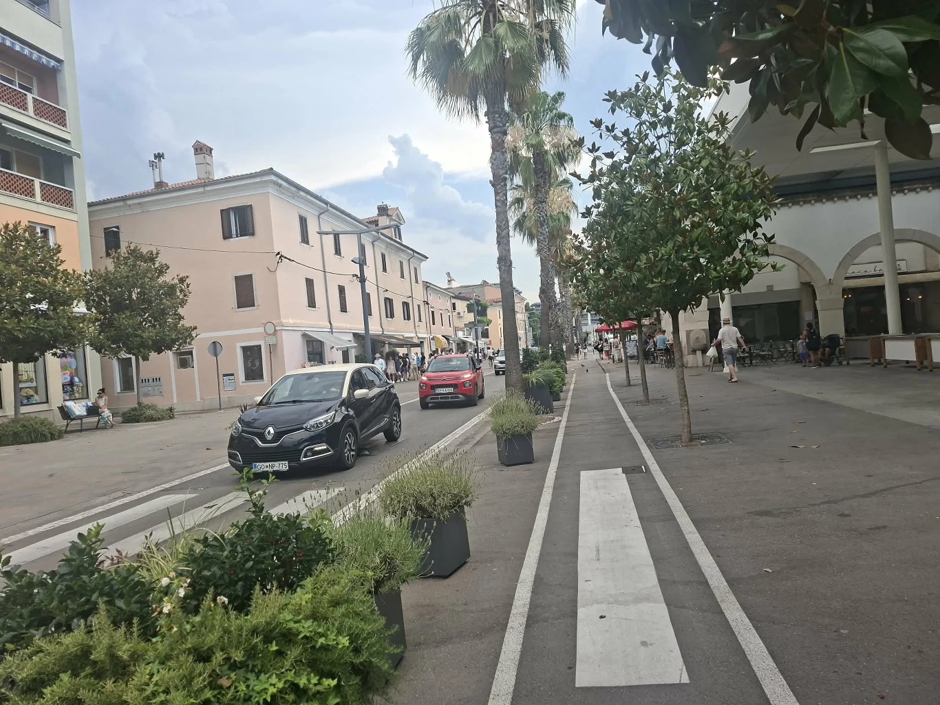 Handlowa promenada w Koprze Ulica z wyznaczoną ścieżką rowerową, rzędem palm oraz kilku zaparkowanymi samochodami przy pastelowych budynkach mieszkalnych, spacerujący przechodnie i widoczne zadaszenie budynku po prawej stronie.