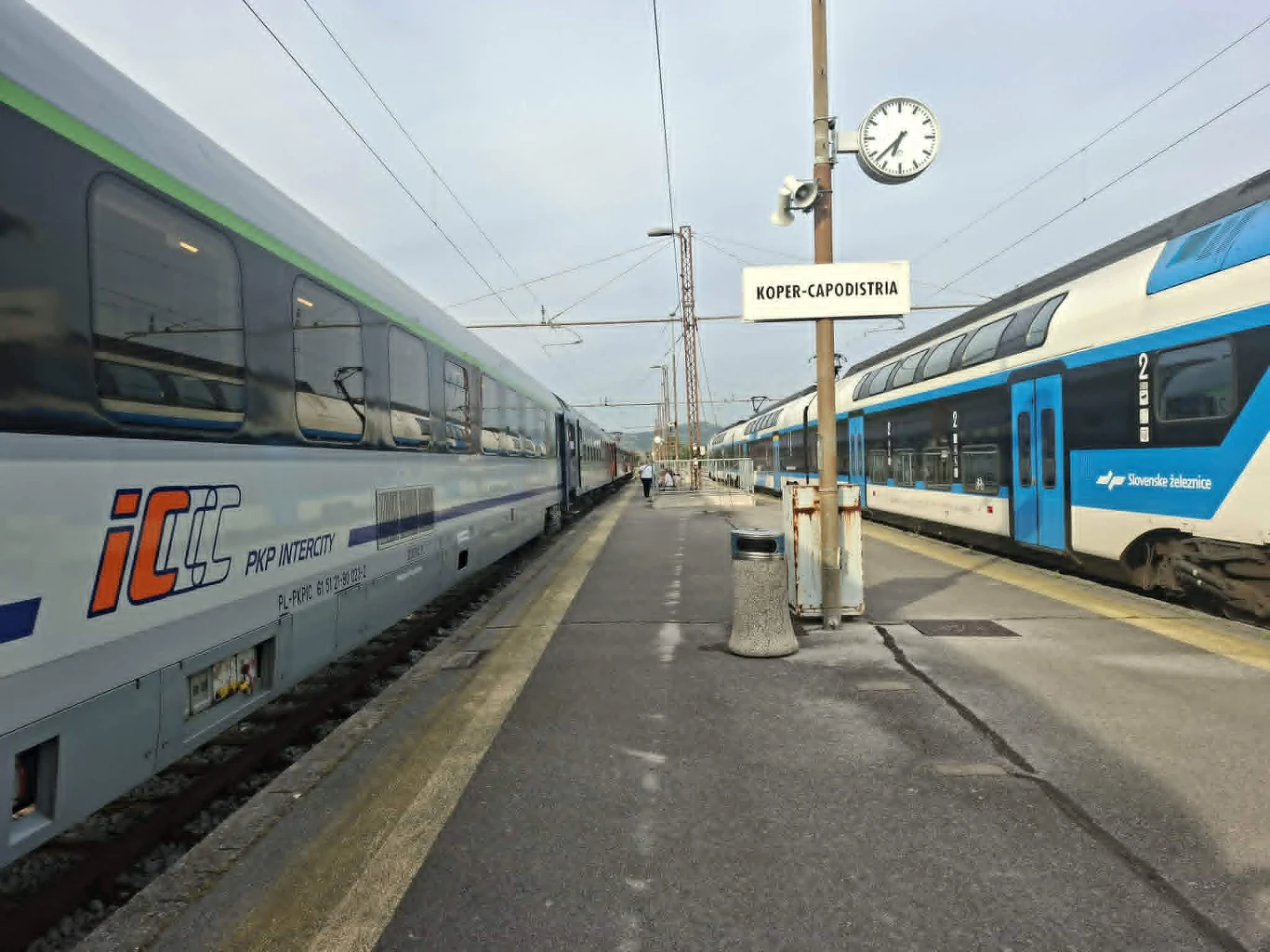 Kolej w Koprze - wagon PKP Intercity doczepiony do węgierskich oraz słoweński, piętrowy zespół trakcyjny Dwa pociągi pasażerskie zatrzymane przy peronie stacji kolejowej Koper-Capodistria, zegar na słupie wskazujący godzinę oraz wyraźnie widoczny znak z nazwą stacji.