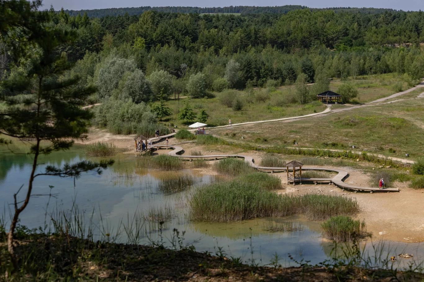 drewniane pomosty nad brzegiem naturalnego zbiornika wodnego z otaczającą je zielenią i lasem w tle, pojedyncze osoby spacerujące po terenie rekreacyjnym