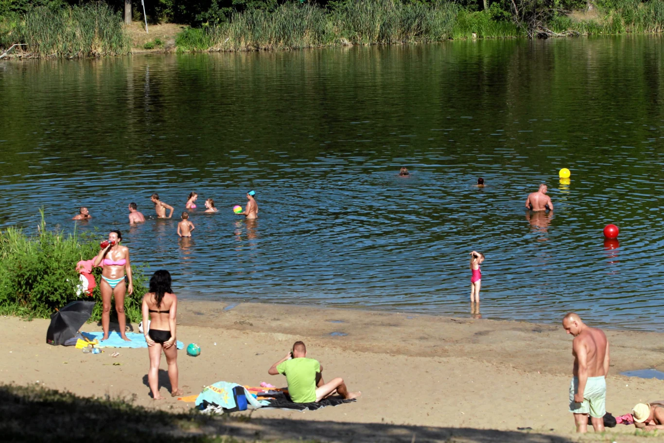 Grupa ludzi wypoczywa nad jeziorem, część osób pływa w wodzie, a inni opalają się i spacerują po piaszczystej plaży.