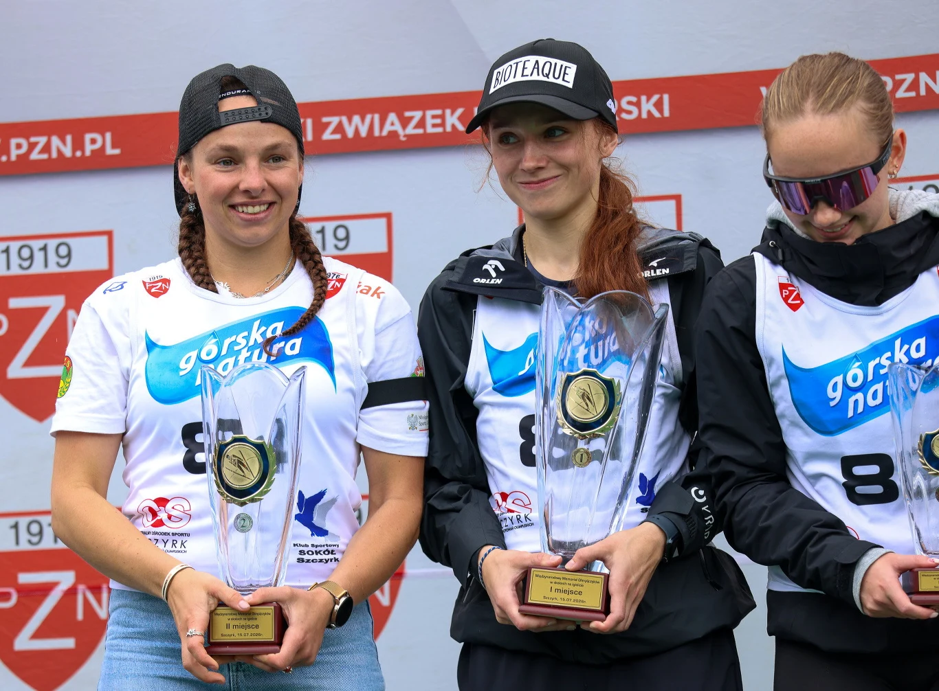 Trzy zawodniczki stojące na podium z pucharami i medalami, ubrane w sportowe stroje z widocznymi numerami oraz nazwą sponsoringu, uśmiechnięte po zakończeniu zawodów.