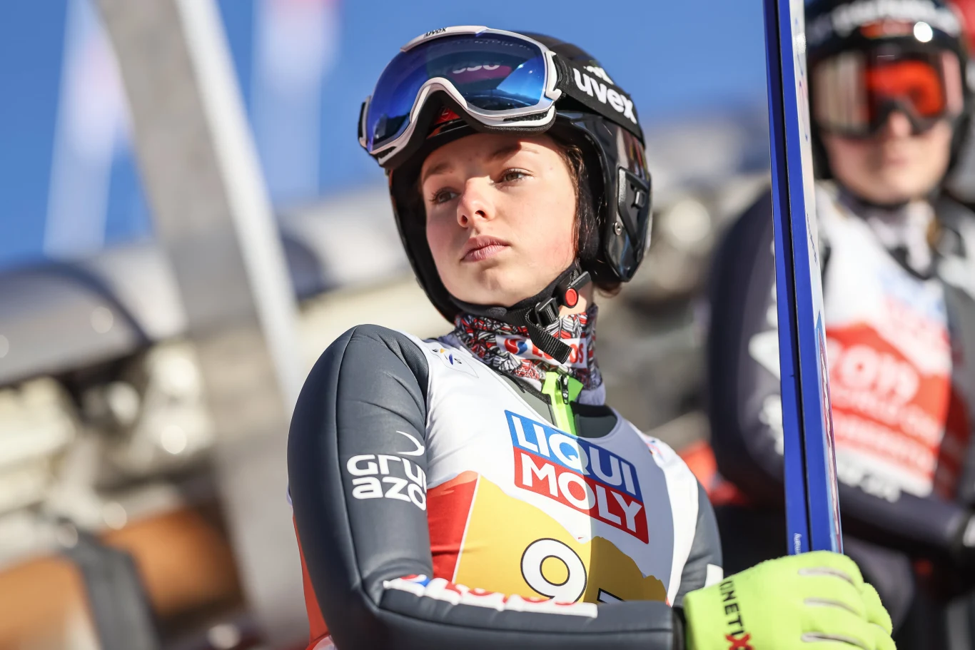 Zawodniczka w kombinezonie sportowym i kasku narciarskim, trzymająca narty i skupiona na starcie, z widoczną reklamą sponsora na stroju.