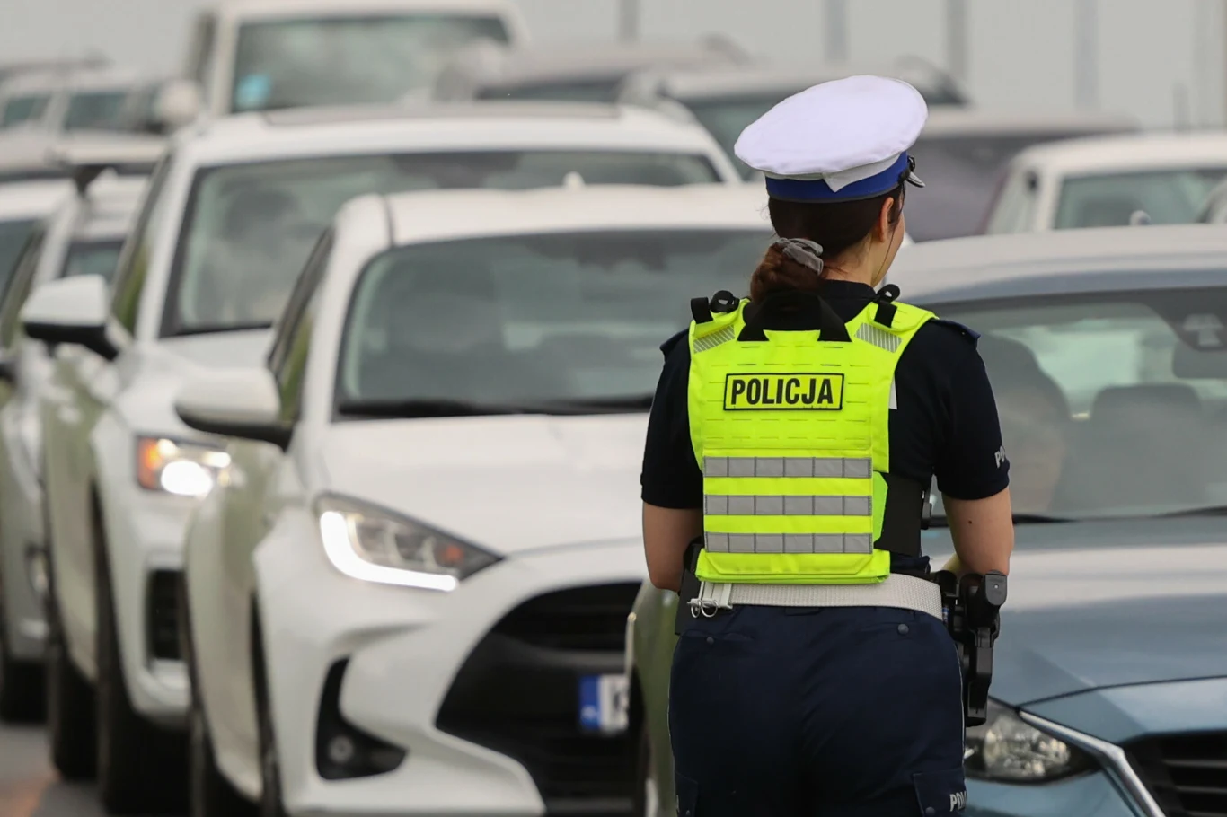 Policjantka w kamizelce odblaskowej i czapce służbowej kontroluje samochody stojące w korku na drodze.
