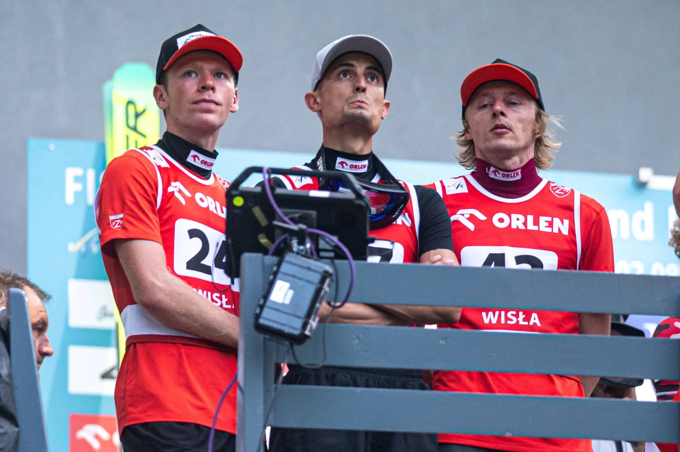Trzech sportowców w czerwonych koszulkach z numerami startowymi i czapkami skupia uwagę na monitorze, prezentując skoncentrowane wyrazy twarzy.