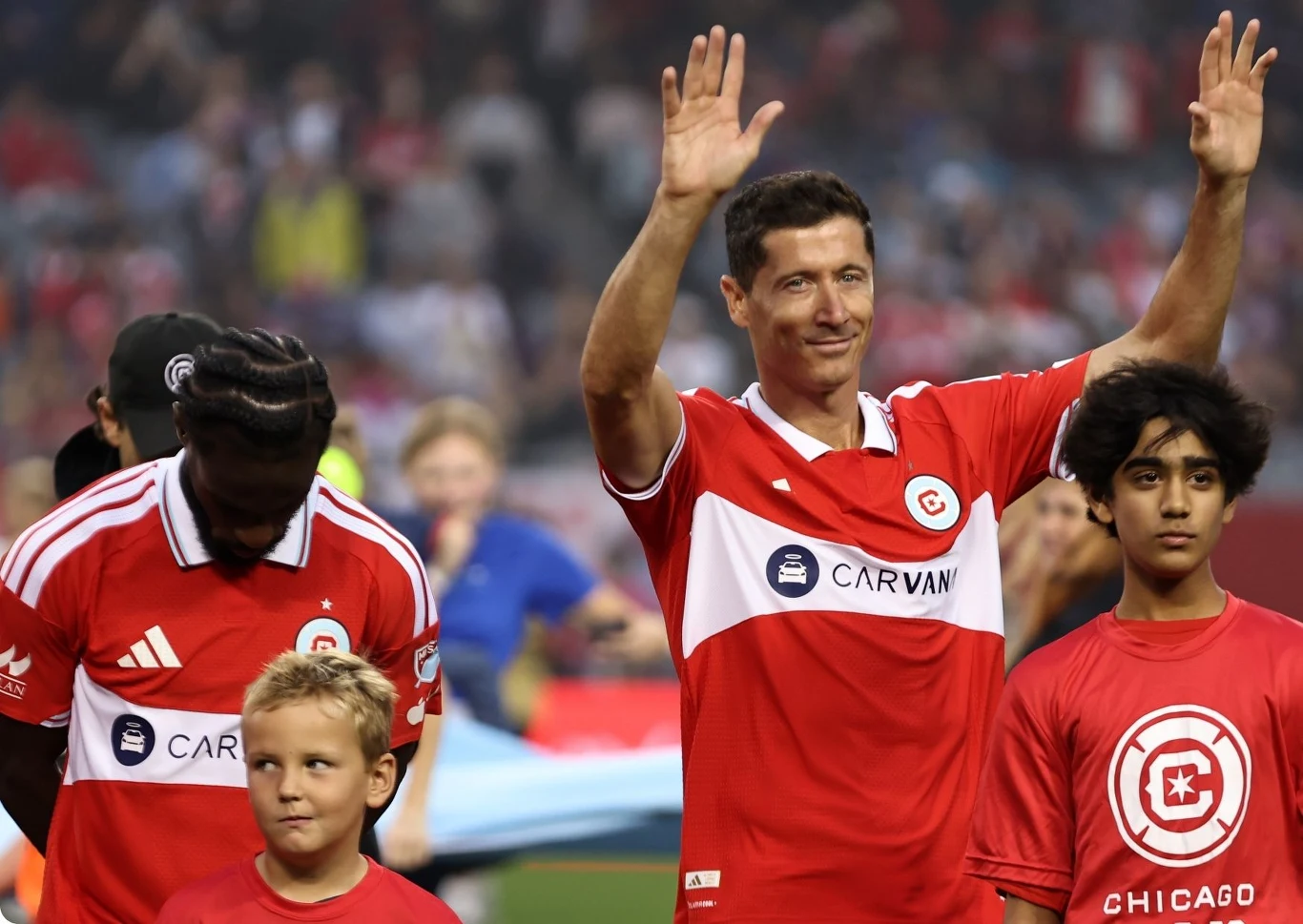 Robert Lewandowski Piłkarz w czerwonej koszulce z podniesionymi rękami stoi w otoczeniu innych zawodników i dzieci na stadionie w trakcie sportowego wydarzenia.