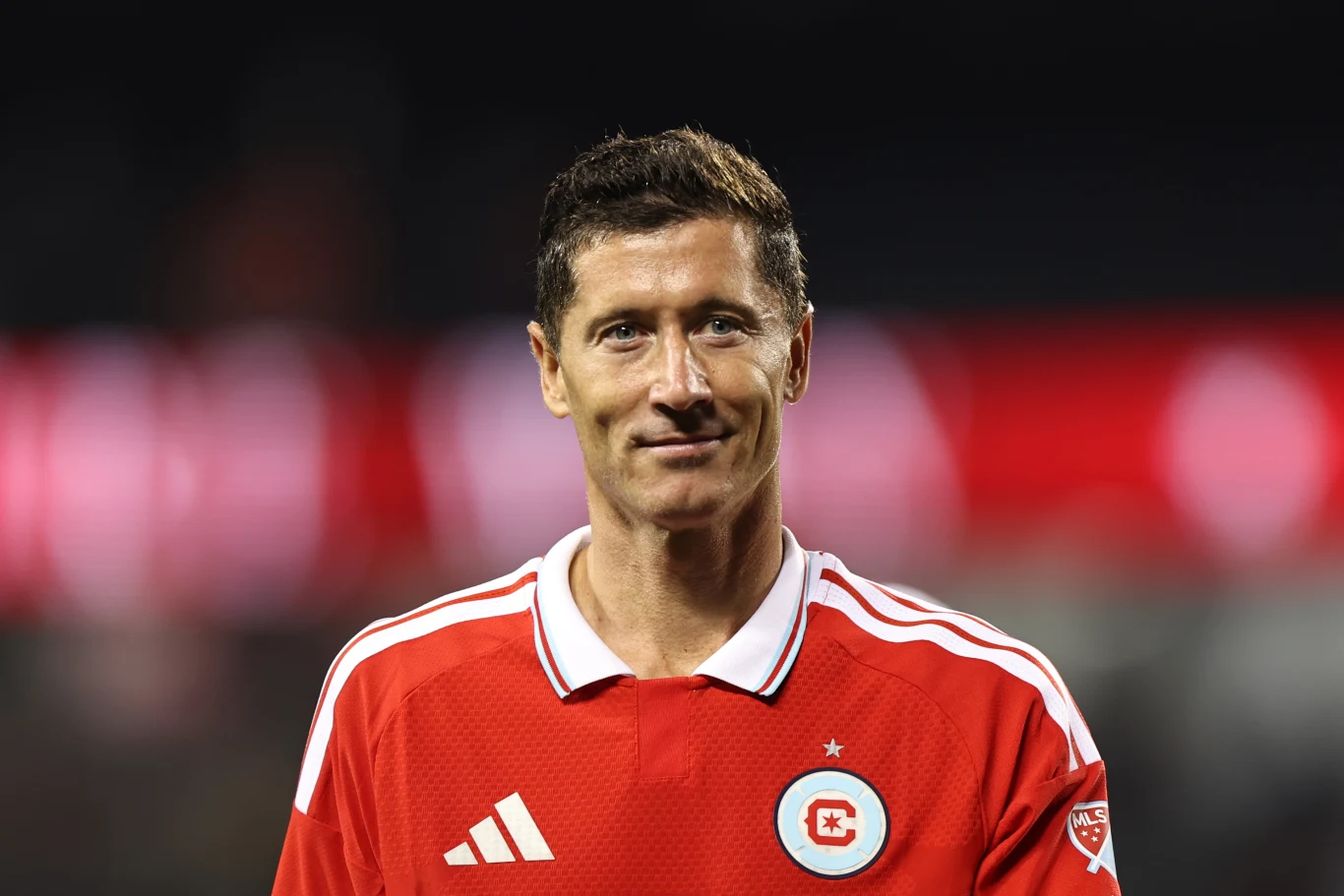 Mężczyzna w czerwonej koszulce sportowej z białymi detalami i emblematem na piersi stoi na tle rozmytego stadionu, patrząc wprost z lekkim uśmiechem.