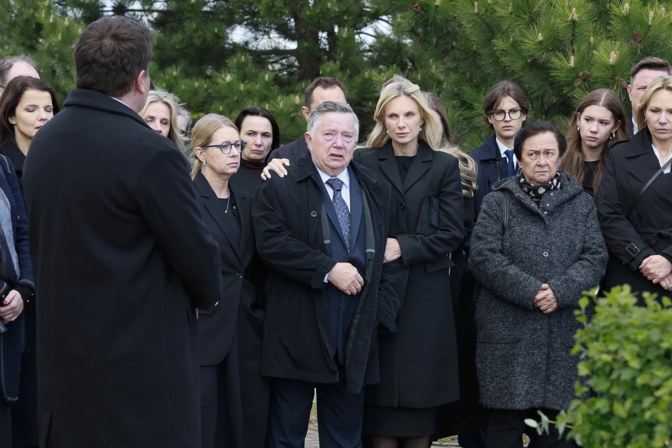 Kadr z pogrzebu Marii Zięby z "Na Wspólnej" Grupa dorosłych osób ubranych na czarno zgromadzona wokół mężczyzny w garniturze, wyrażająca smutek i zadumę, otoczona zielenią drzew.
