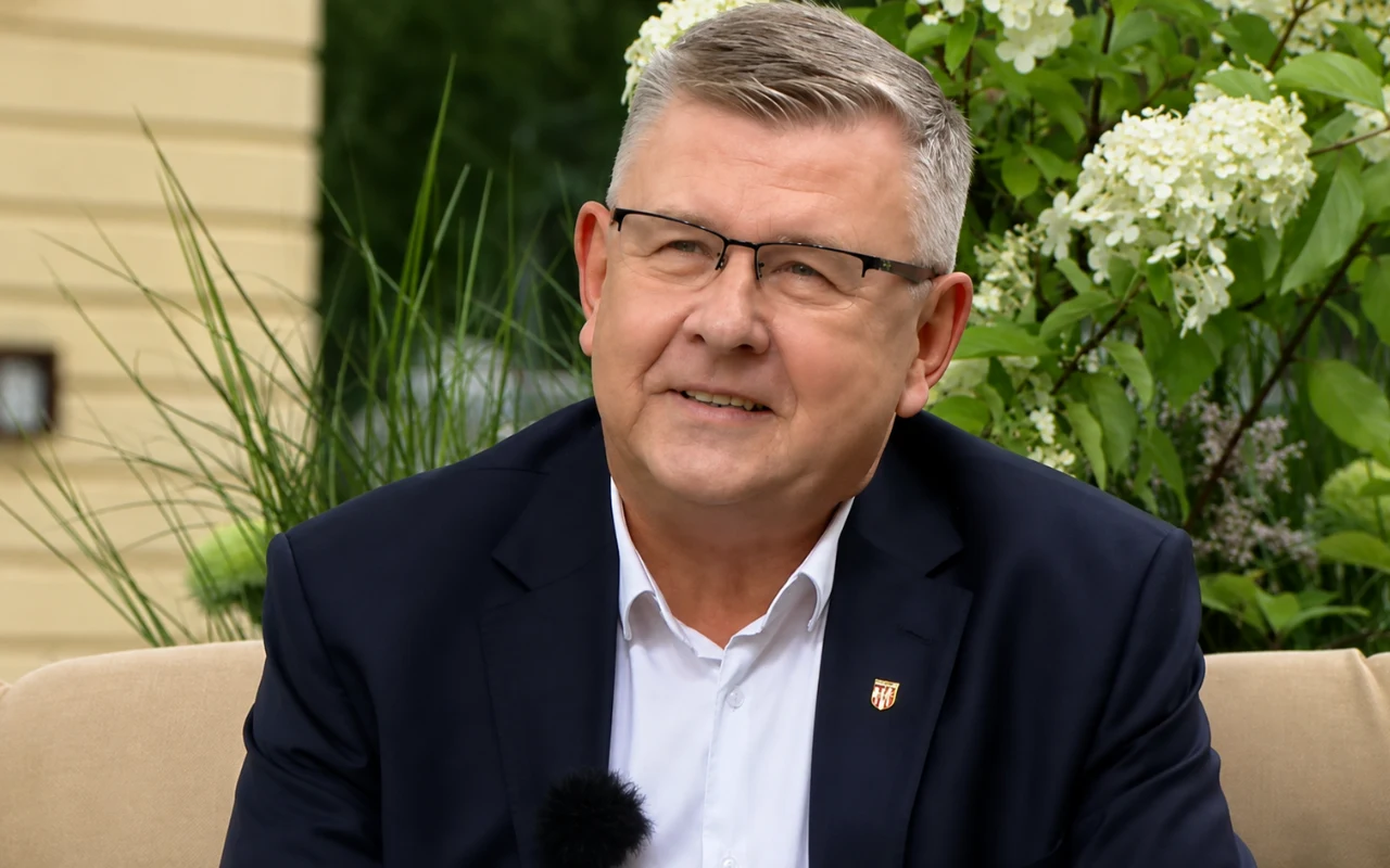 Witold Kozłowski Witold Kozłowski w okularach i granatowej marynarce siedzi na jasnej sofie w ogrodzie na tle zieleni i białych kwiatów.