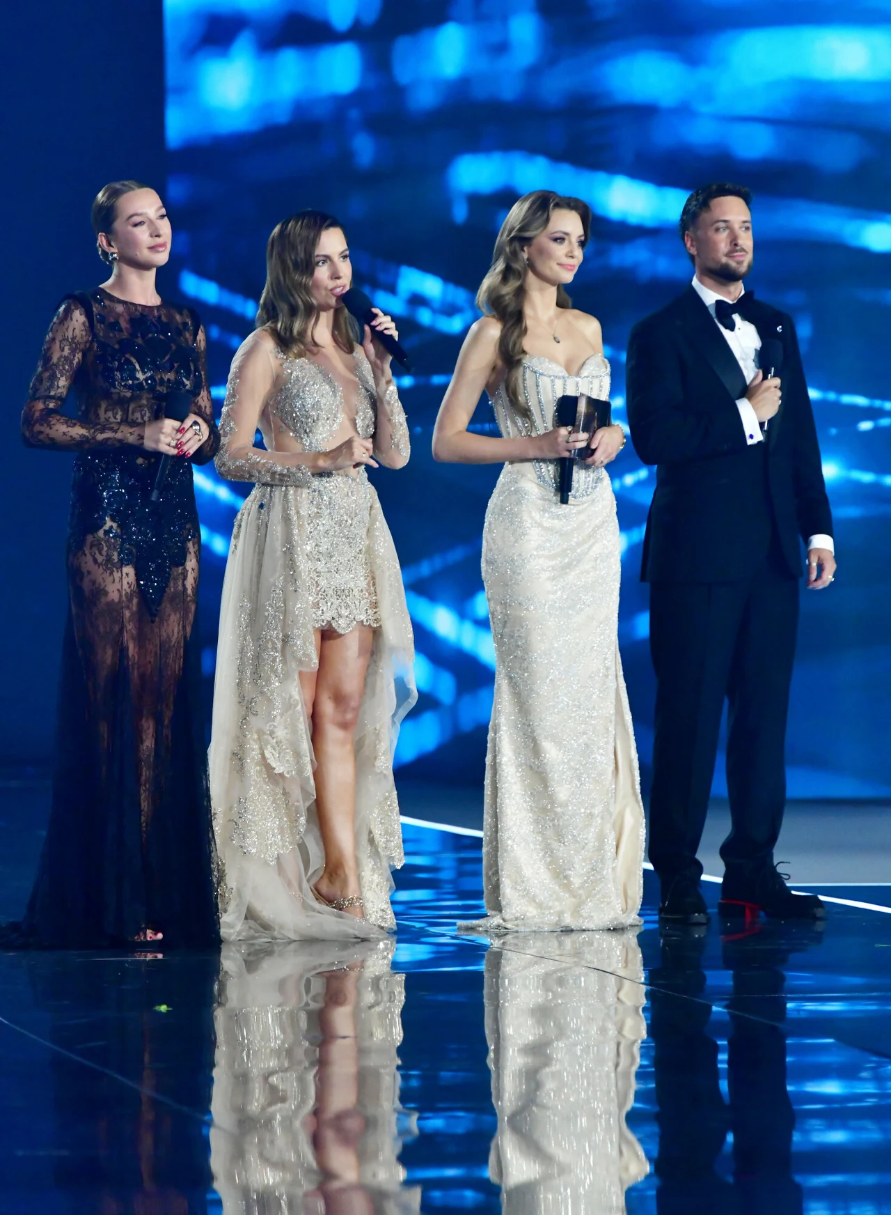 Trzy kobiety w eleganckich wieczorowych sukniach oraz mężczyzna w smokingu stoją obok siebie na scenie, trzymając mikrofony na tle niebieskiego, futurystycznego podświetlenia.