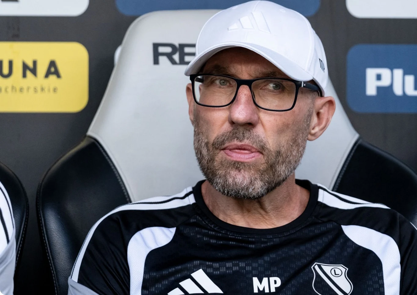 Marek Papszun Mężczyzna w białej czapce z daszkiem i okularach siedzi na sportowym fotelu, ubrany w czarno-biały strój z logo klubu piłkarskiego Legia Warszawa.