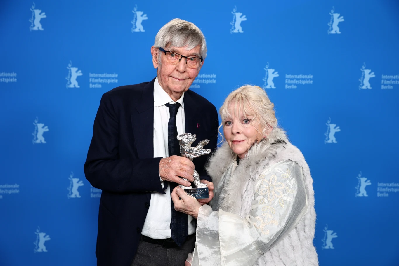 Tom Courtenay i Anna Calder-Marshall z nagrodą na festiwalu w Berlinie za role w "Daleko od brzegu" Dwoje elegancko ubranych seniorów pozuje razem na tle niebieskiej ścianki z logo festiwalu filmowego, trzymając w rękach srebrną statuetkę z motywem niedźwiedzia.